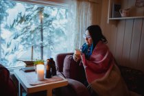 Вид збоку молодої жінки в окулярах під ковдрою відпочиває на дивані з чашкою біля вікна з видом на ліс у снігу — стокове фото