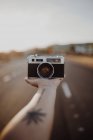 Mão de colheita de pessoa com câmera vintage na palma da mão entre a rota de asfalto no fundo embaçado — Fotografia de Stock