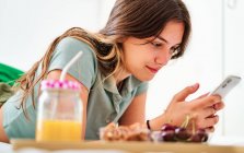 Vista lateral da jovem estudante navegando redes sociais no celular perto da mesa com frutas frescas e suco enquanto passa a manhã em casa — Fotografia de Stock