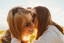 Feliz morena dando un beso a la rubia mejor amiga en gafas de sol sentada en la luz dorada del atardecer en la naturaleza - foto de stock