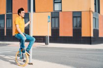 Вид сбоку на все тело молодого парня в повседневной одежде, сидящего на велосипеде на современной городской улице с разноцветным зданием — стоковое фото