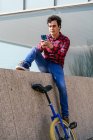 Full body of young guy in checkered shirt and jeans sitting on stone fence near unicycle and browsing mobile phone during free time in city — Stock Photo