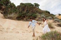 Delighted multiracial bridge and groom holding hands and running along sandy hill on wedding day in nature — Stock Photo