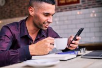 Hombre hispano feliz con camisa violeta disfrutando de una bebida caliente y navegando por las redes sociales en el teléfono celular mientras toma un descanso durante el trabajo en la cafetería - foto de stock