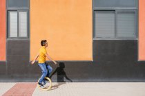 Повний вид з боку тіла сучасного молодого чоловіка в яскраво-помаранчевій сорочці і джинсах, що слухають музику через бездротові навушники під час їзди на одноколісному велосипеді на тротуарі біля міської будівлі — стокове фото
