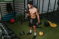 Treinador masculino jovem muscular com torso nu levantando halteres pesados durante o treinamento no ginásio — Fotografia de Stock