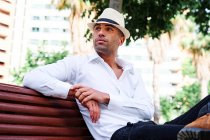 Confiante bonito jovem hispânico masculino em roupas elegantes e chapéu sentado no banco e olhando para longe, enquanto descansa na rua da cidade — Fotografia de Stock