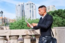 Vista lateral del joven gerente ejecutivo hispano enfocado en traje elegante con información de lectura de bolsos en la tableta mientras está parado cerca de la barandilla en la terraza urbana - foto de stock