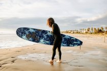 Vista lateral del surfista vestido con traje de neopreno caminando con tabla de surf hacia el agua para coger una ola en la playa durante el amanecer - foto de stock
