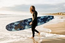 Vista lateral do surfista vestido de fato de mergulho andando com prancha de surf em direção à água para pegar uma onda na praia durante o nascer do sol — Fotografia de Stock