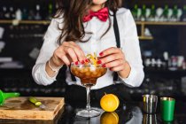 Cropped unrecognizable female barkeeper in stylish outfit decorating cocktail with lemon peel while standing at counter in modern bar — Stock Photo