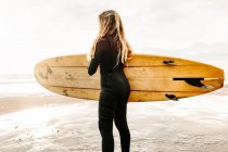 Vista lateral da surfista mulher vestida de fato de mergulho de pé olhando para longe com a prancha de surf na praia durante o nascer do sol no fundo — Fotografia de Stock