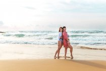 Full body of cheerful girlfriends in swimwear hugging each other while standing on sandy beach washed by waving sea — Stock Photo
