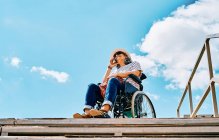 Mature disabled female sitting in wheelchair and talking on mobile phone near stairway against blue sky in city — Stock Photo