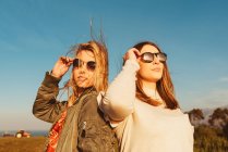 Young close female friends in stylish clothes standing together on meadow in mountains looking away in golden light — Stock Photo