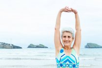 Antiguo pensionista femenina de pelo gris activa en traje de baño elegante estirando los brazos y los hombros mientras hace ejercicios en la playa contra el océano ondulante - foto de stock