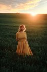 Junge Frau im Vintage-Kleid schaut nachdenklich weg, während sie allein auf der Wiese bei Sonnenuntergang am Sommerabend auf dem Land steht — Stockfoto