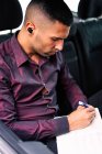 Homme d'affaires hispanique sérieux avec de vrais écouteurs sans fil lisant des notes dans un ordinateur portable assis sur le siège arrière de la voiture et se rendant au travail — Photo de stock