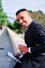 Side view of happy young Hispanic man in elegant formal suit drinking takeaway beverage and browsing tablet while having free time and resting in park in sunny summer day — Stock Photo