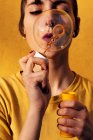 Modern female with piercing blowing soap bubbles with closed eyes at camera on sunny day against yellow wall — Stock Photo