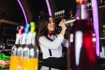 Felice barista donna in elegante vestito agitando agitatore di metallo mentre si prepara cocktail in piedi al bancone nel bar moderno — Foto stock