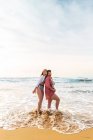 Full body of cheerful girlfriends with mouth opened hugging while standing looking at camera on sandy beach washed by waving sea — Stock Photo