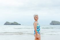 Seitenansicht einer gealterten grauhaarigen Frau in stylischer Badebekleidung, die lächelt und in die Kamera schaut, während sie an einem Sommertag am Strand vor dem wogenden Meer steht — Stockfoto