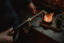 Неузнаваемый ювелир плавит металл для ювелирных изделий с паяльной лампой, стоя возле верстака в мастерской — стоковое фото