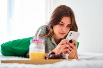 Jovem estudante navegando redes sociais no celular perto da mesa com frutas frescas e suco enquanto passa a manhã em casa — Fotografia de Stock