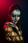 Modisches junges weibliches Modell mit Lichtprojektion in Form orientalischer Hieroglyphen, die in einem dunklen Atelier mit roter Beleuchtung nach unten schauen — Stockfoto