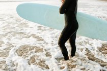 Vista lateral del surfista irreconocible vestido con traje de neopreno de pie con la tabla de surf en el agua esperando para coger una ola en la playa durante el amanecer - foto de stock