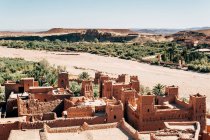 Ancient buildings of brown stone among green tropical plants and desert with clear blue sky on background in Morocco — Stock Photo
