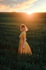 Young female in vintage dress looking away thoughtfully while standing alone in grassy field at sunset time in summer evening in countryside — Stock Photo