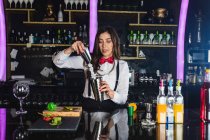 Женщина-бармен в стильном наряде добавляет кубики льда в шейкер во время приготовления коктейля стоя у стойки в современном баре — стоковое фото