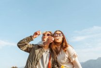 Chiudi amiche donne in occhiali da sole che soffiano bolle di sapone insieme in piedi in abbraccio sul prato in montagna — Foto stock