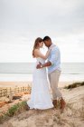 Vista lateral de amante pareja multirracial de recién casados abrazándose mientras está de pie en la colina de arena en el fondo del mar en el día de la boda - foto de stock