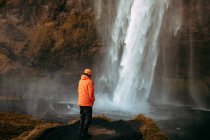 Visão traseira do turista jovem em desgaste de inverno olhando para cascata e rio de montanha entre colina de pedra — Fotografia de Stock