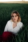 Спокійна молода жінка в ретро-стилі біла блузка сидить серед високої зеленої трави і дивиться на камеру, відпочиваючи в літній вечір у сільській місцевості — стокове фото