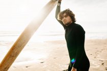 Vue latérale de surfeur souriant homme vêtu d'une combinaison de plongée debout regardant la caméra avec la planche de surf sur la plage pendant le lever du soleil en arrière-plan — Photo de stock
