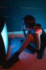 Vista lateral do modelo feminino irreconhecível em vestido preto sentado no chão perto de brilhar televisão antiga no estúdio escuro — Fotografia de Stock