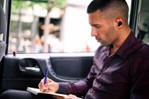 Homem empreendedor hispânico focado sentado no assento do passageiro do carro e escrevendo no planejador enquanto viaja para o trabalho — Fotografia de Stock