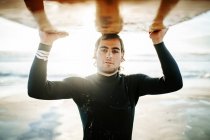 Portrait of young surfer man dressed in wetsuit standing looking at camera on the beach with the surfboard above head during sunrise — Stock Photo