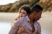 Loving woman hugging black man from behind while spending summer day together on seashore — Stock Photo