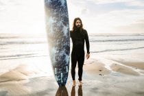 Серфер чоловік з довгим волоссям і бородою, одягнений у гідрокостюм, стоїть на камеру з дошкою для серфінгу на пляжі під час сходу сонця на задньому плані — стокове фото