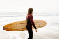 Side view of surfer man dressed in wetsuit walking looking away with surfboard towards the water to catch a wave on the beach during sunrise — Stock Photo