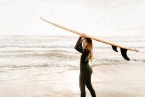 Vista lateral de la surfista vestida con traje de neopreno de pie mirando a la cámara mientras sostiene la tabla de surf en la cabeza en la playa durante el amanecer en el fondo - foto de stock