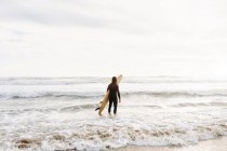 Вид ззаду на невпізнаваного серфінгового чоловіка, одягненого в гідрокостюм, що стоїть, дивлячись з дошкою для серфінгу до води, щоб зловити хвилю на пляжі під час сходу сонця — стокове фото