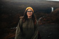 Junger Tourist mit Brille und Hut mit Piercing blickt auf einem Hügel nahe Wasserfall und Gebirgsfluss in die Kamera — Stockfoto