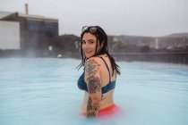 Боковой вид на молодого татуированного хипстера в купальнике, позирующего в синей воде между скалами — стоковое фото