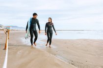 Feliz casal surfista vestido com fatos de mergulho andando com pranchas de surf na costa da praia de areia olhando um para o outro — Fotografia de Stock